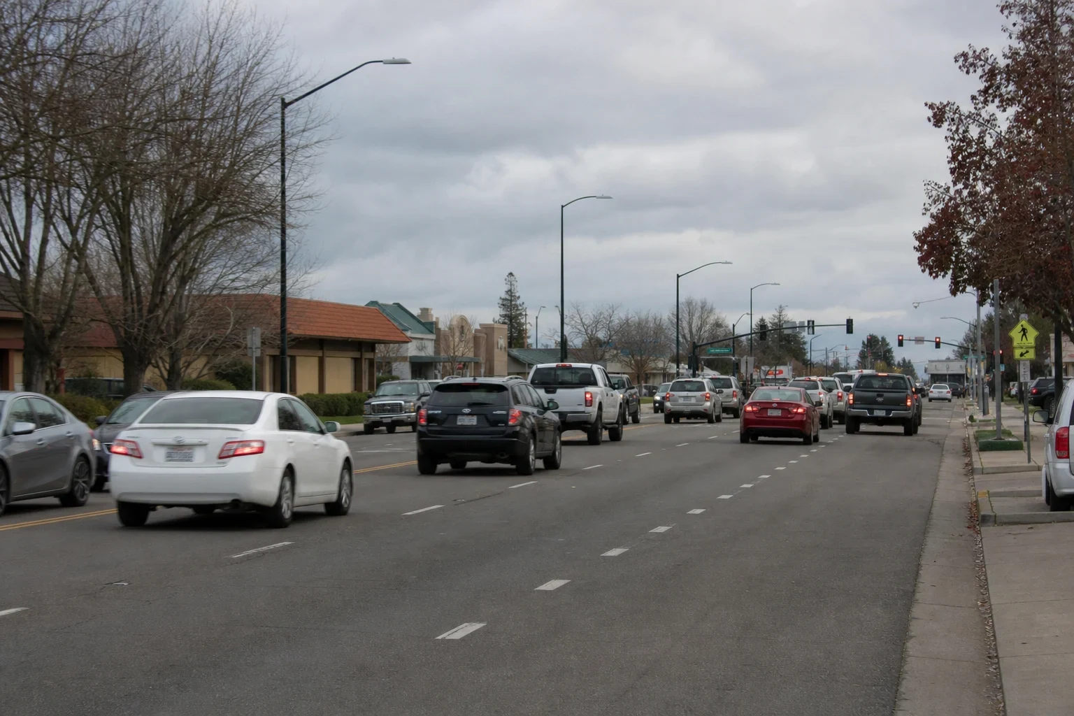 Multi-lane local street with everyday traffic under overcast skies, bare trees, streetlights, traffic signals and low tile-roofed commercial buildings
